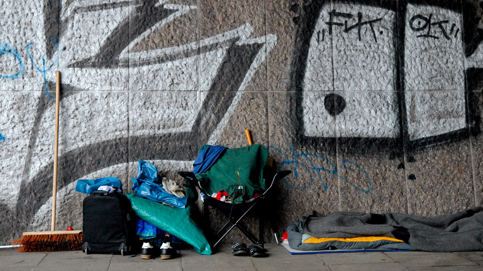 Aufgeräumte, saubere Schlafstätte eines Obdachlosen mit Besen, Stuhl, Schuhen, Schlafsack vor einer Mauer mit Graffiti